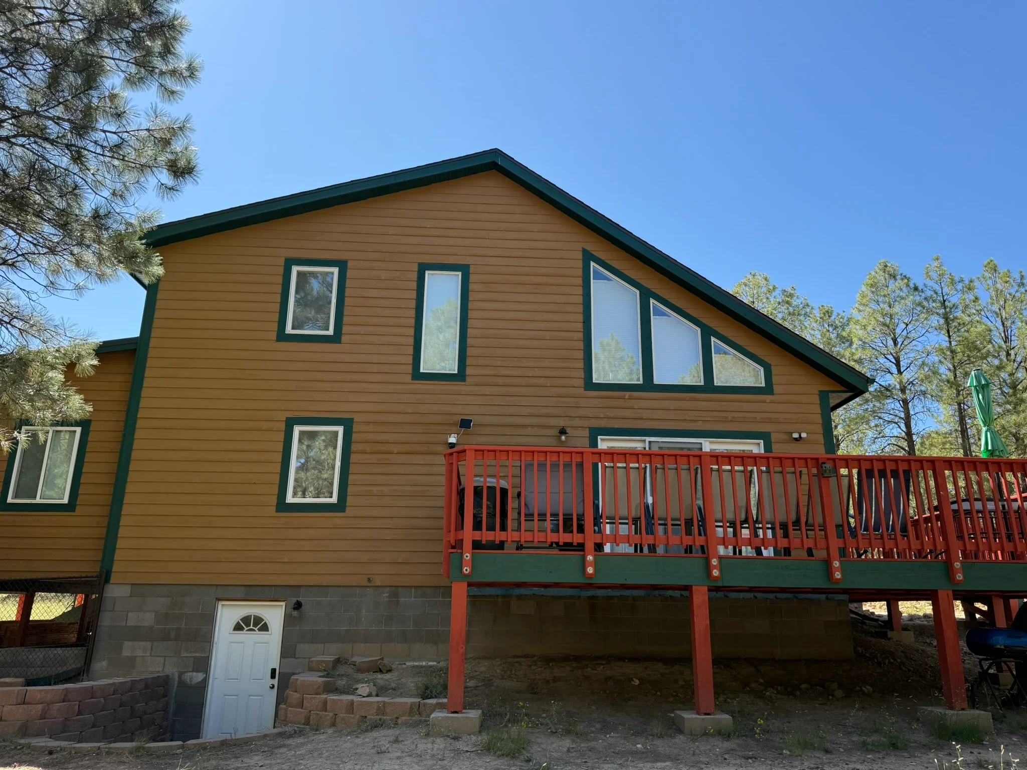 After - Two-story home with golden-yellow lap siding, dark green trim, white window frames, and a red deck railing surrounded by pine trees by Blue J Paint & Stain, Kingman AZ