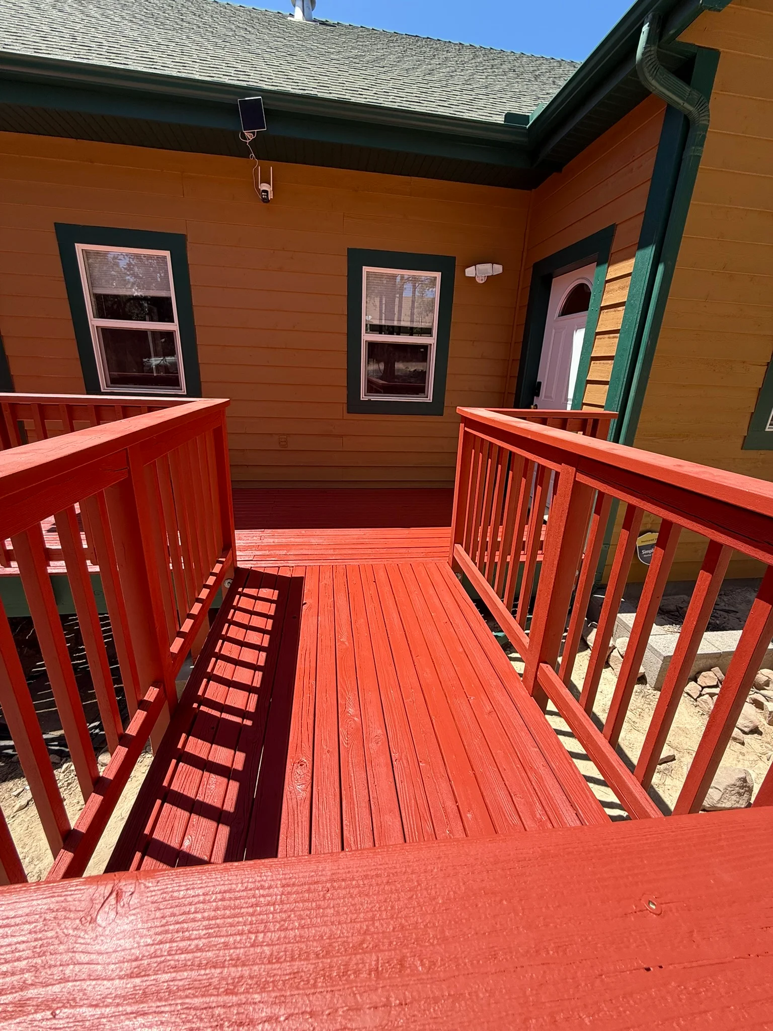 Red painted deck with railings, orange wood siding, dark-trimmed windows, and green entry door